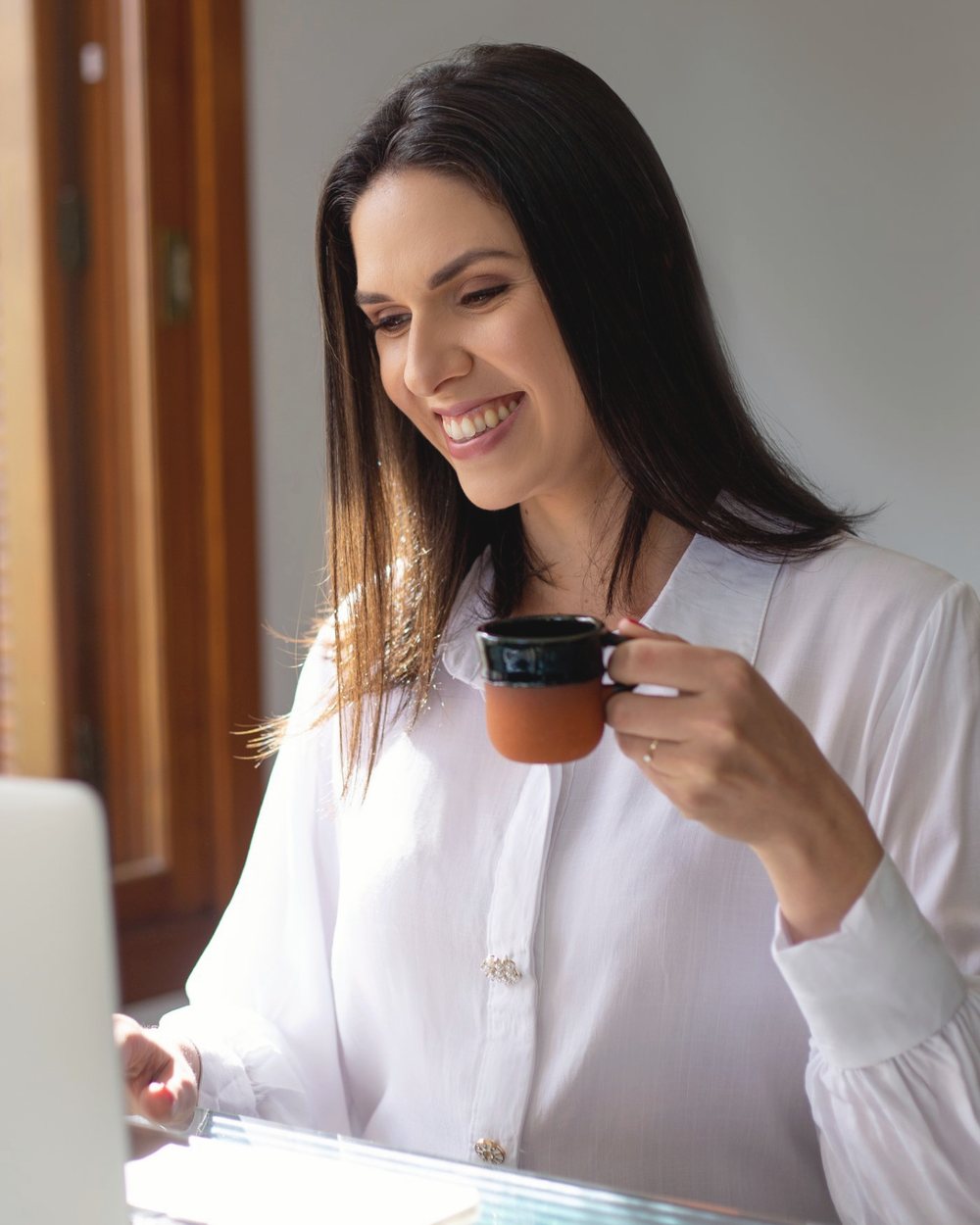 Daiana Prestes com uma xícara de café, sorrindo diante do notebook